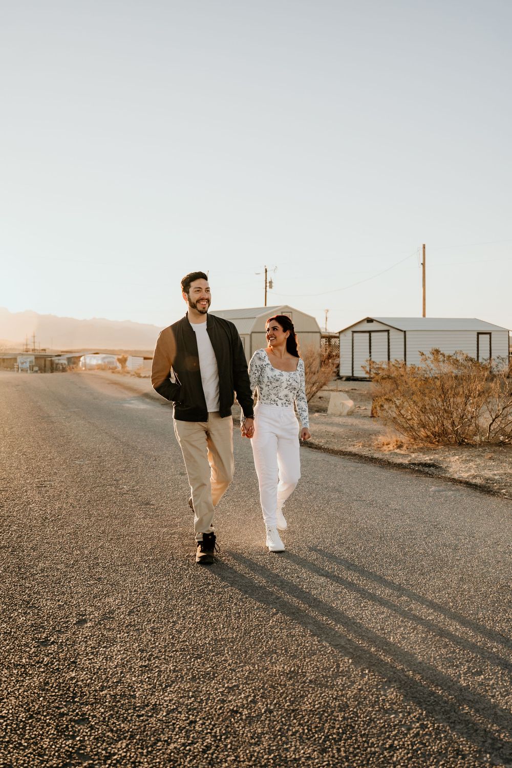 Couple walking together on a desert road at sunset with mobile homes in the background.