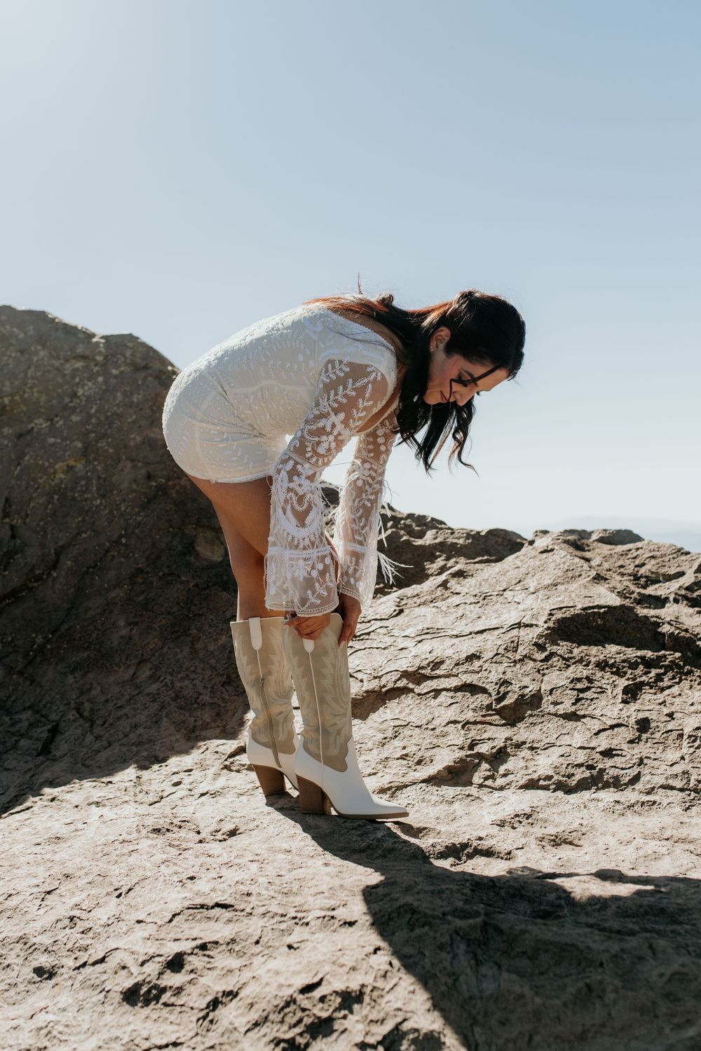 Person in white lace dress adjusting boots on rocky terrain.