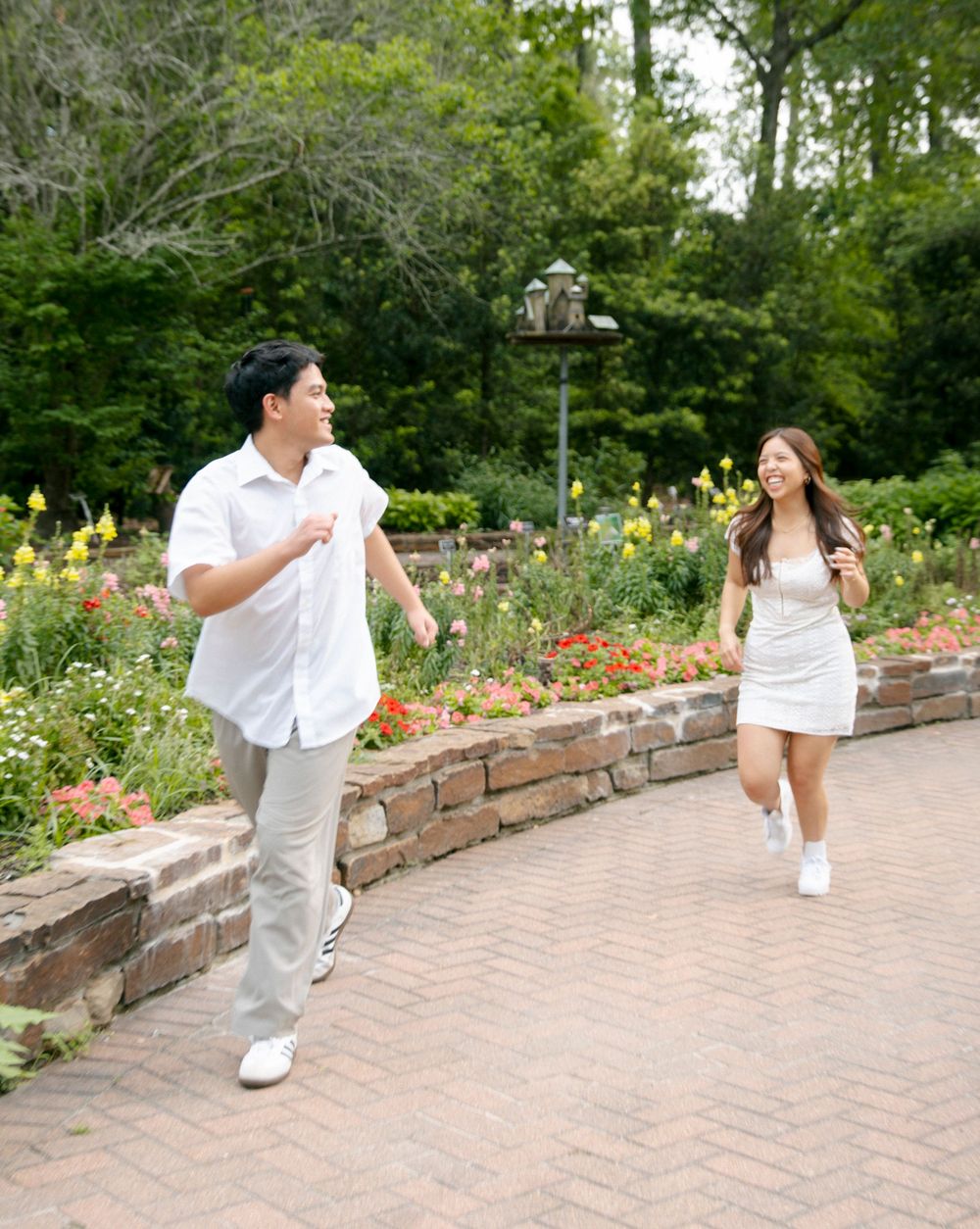 A couple in white outfits shares a playful moment walking along a brick pathway in a garden with blooming flowers.