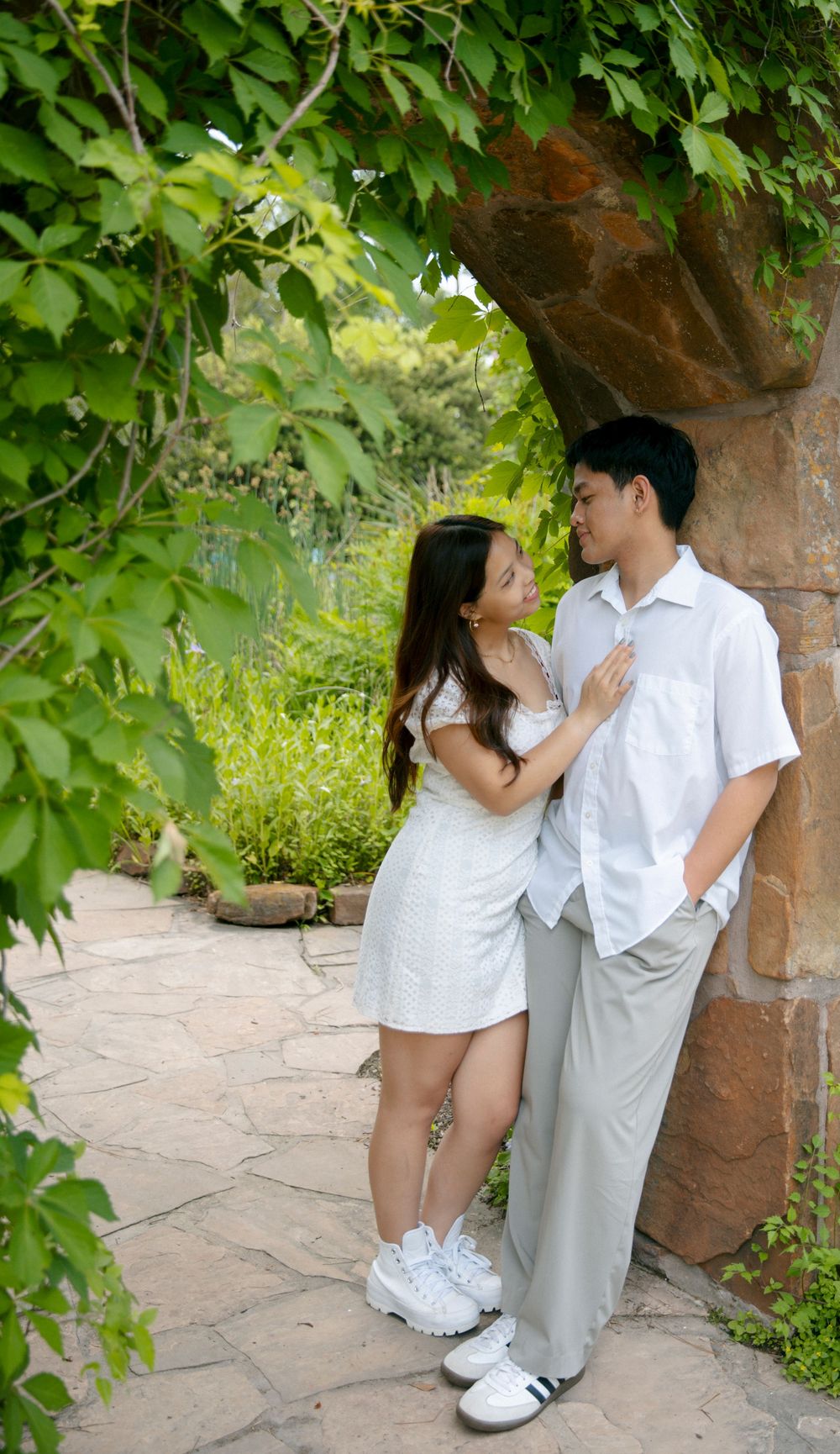 A romantic couple in white outfits share a tender moment under a stone archway surrounded by green foliage.