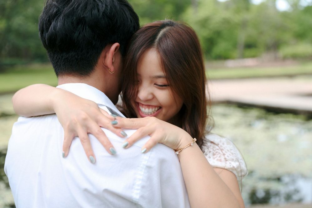 A couple in white wedding attire embraces while standing near a tranquil pond in a garden setting.