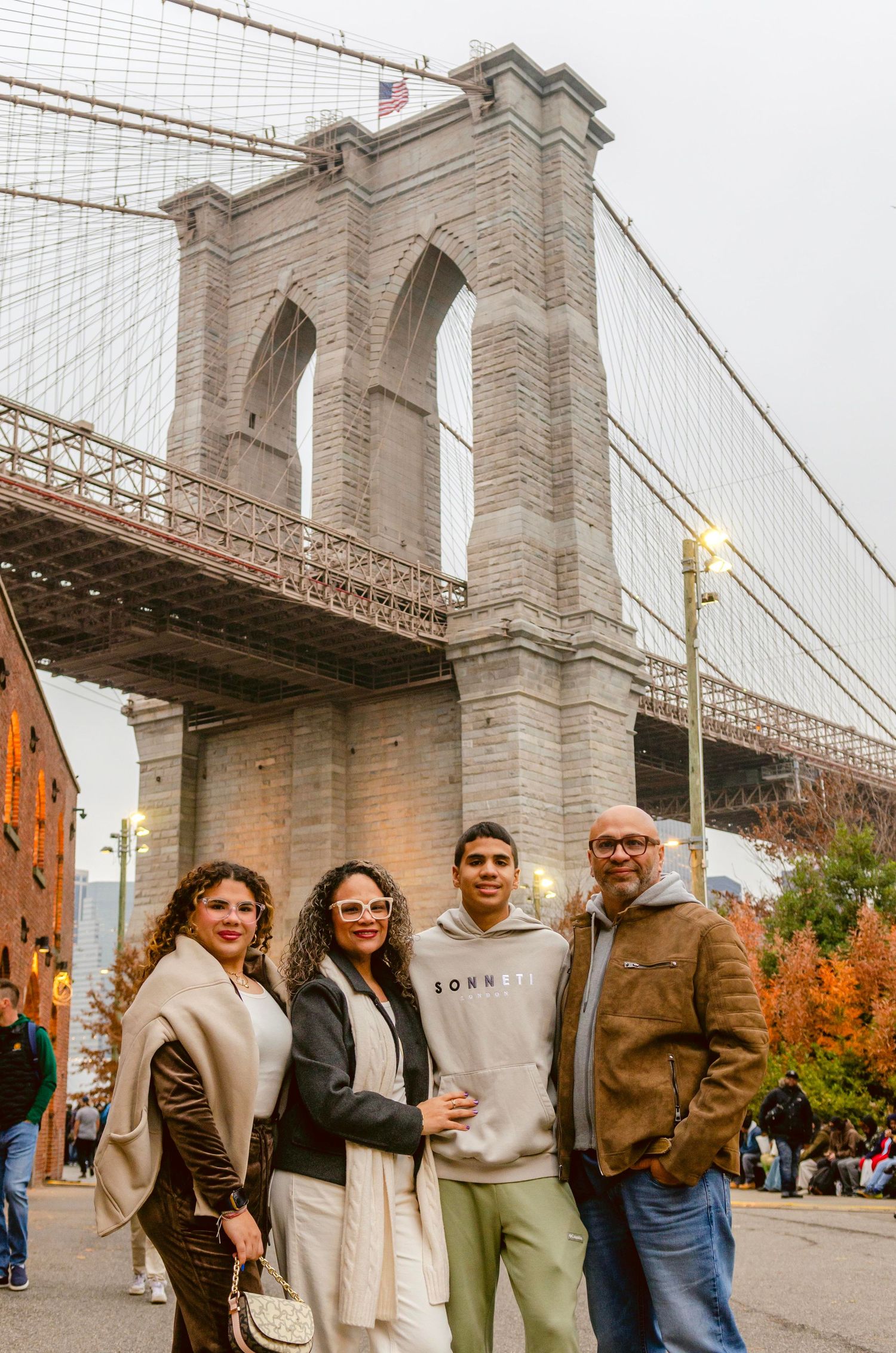 A group stands together beneath the iconic Brooklyn Bridge on an autumn day with red brick buildings in the background.