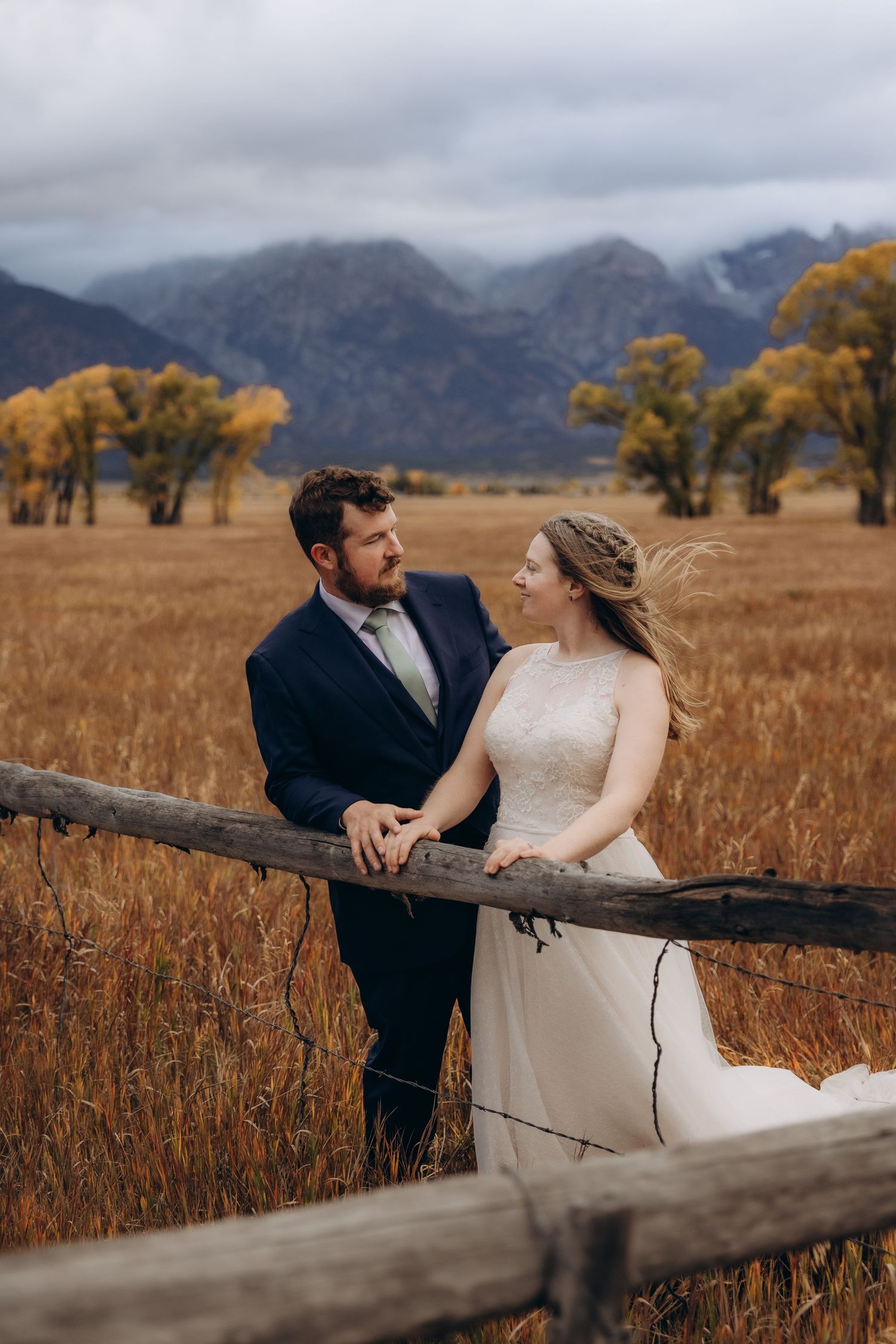 A couple in wedding attire poses together near a rustic wooden fence with mountains and autumn foliage in the background.