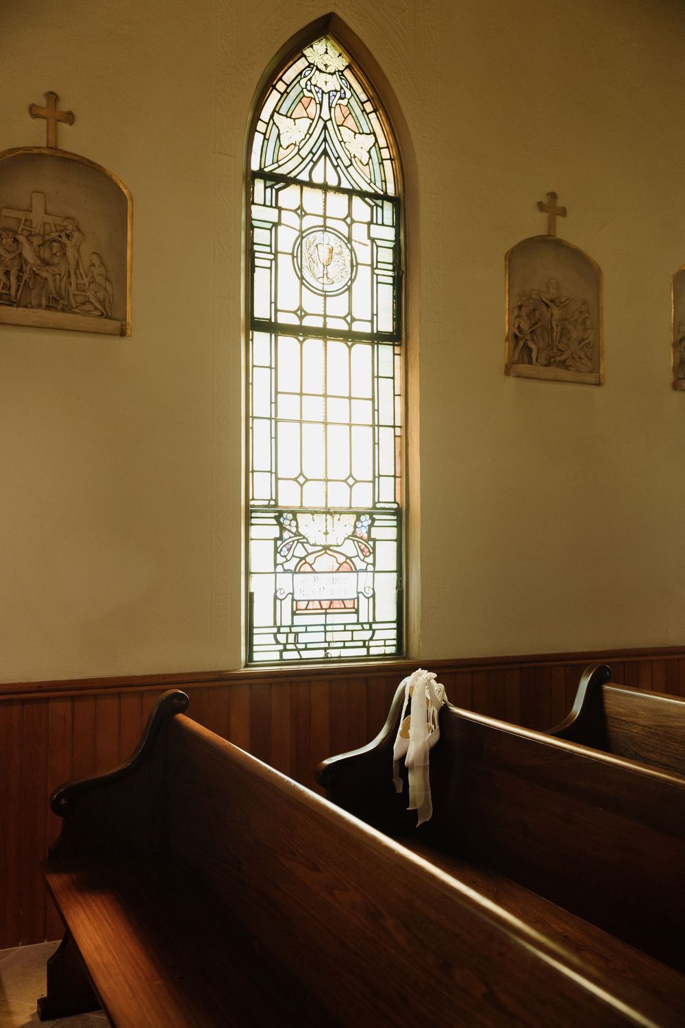 A stained glass church window illuminates wooden pews while a white cloth drapes over one of the benches.