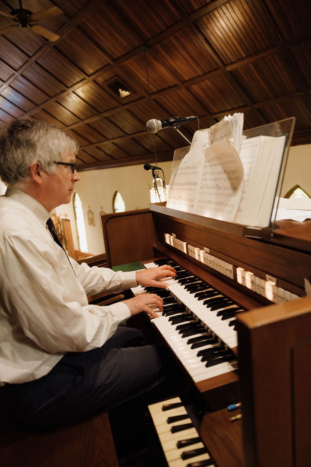 A pianist plays an upright piano in a church or chapel setting with warm wooden interior lighting.