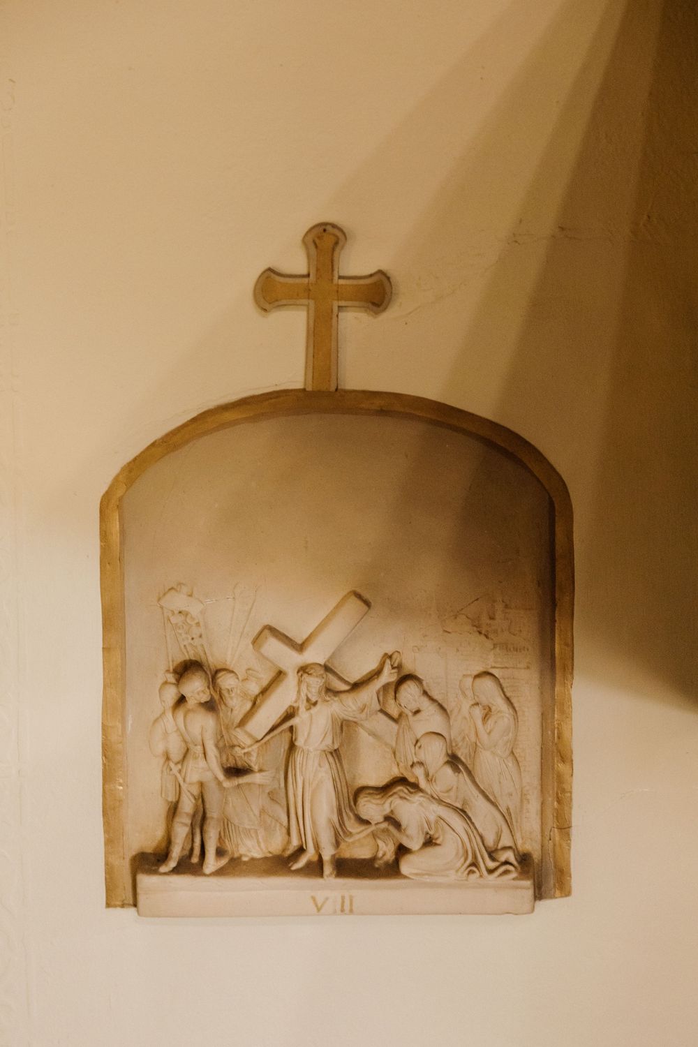 A sepia-toned religious wall relief depicting the Stations of the Cross with a cross mounted above in an arched alcove.