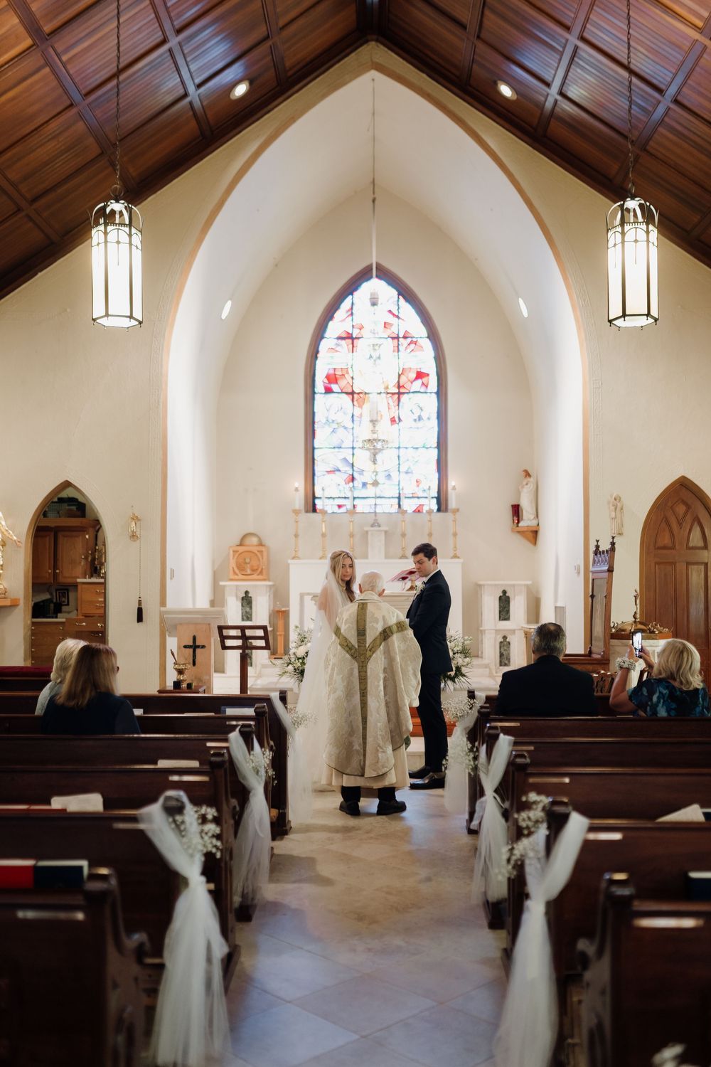 Interior of a traditional church with vaulted ceiling, stained glass window, and hanging lanterns during a ceremony.