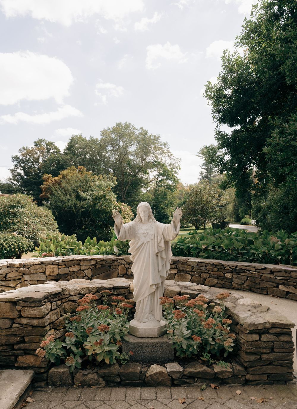 Stone wall garden path with religious statue and flowering plants in a peaceful outdoor setting.