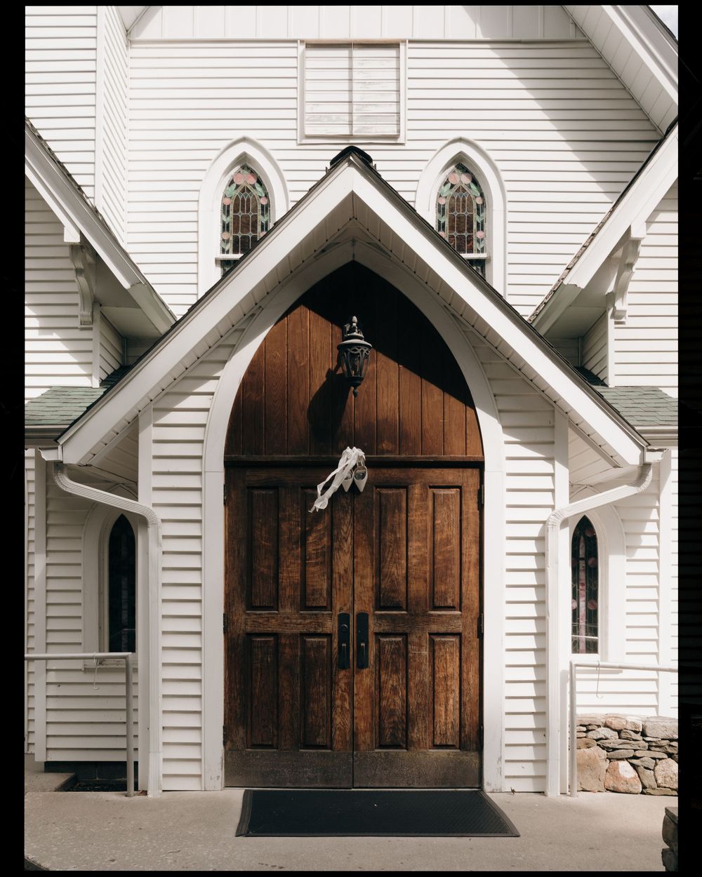 A gothic-style wooden church door with pointed arch and white trim features a decorative metal handle on a sunny day.