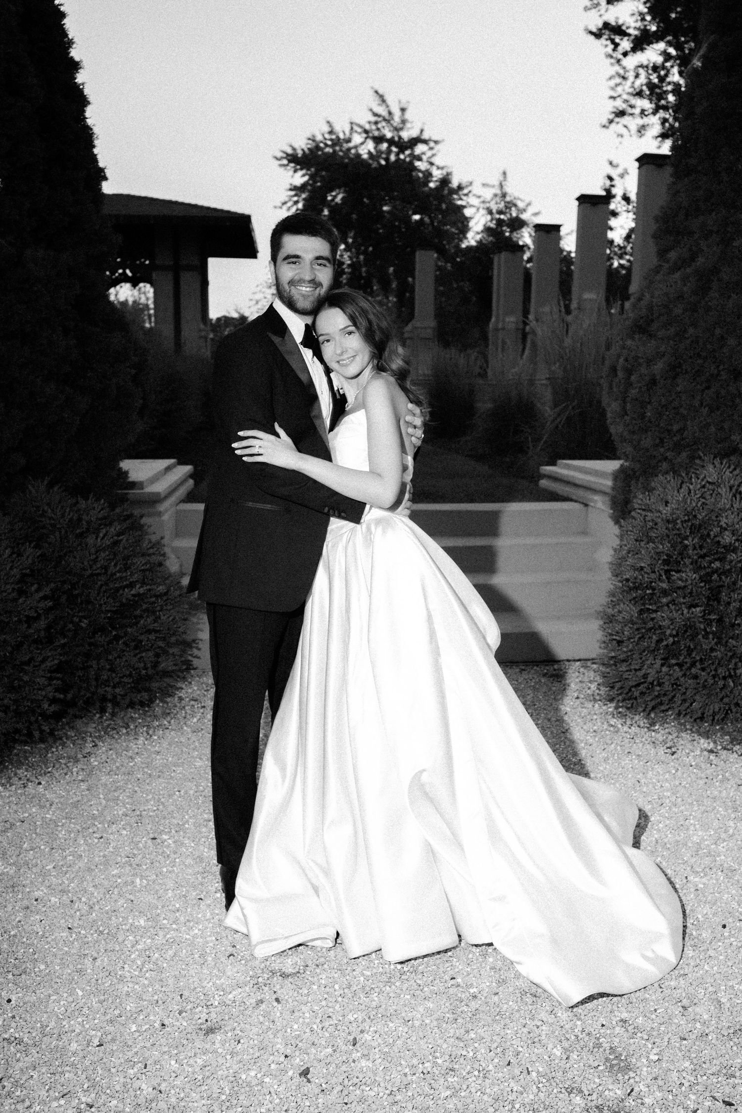 A bride and groom embrace in a formal garden setting during their black and white wedding photoshoot.