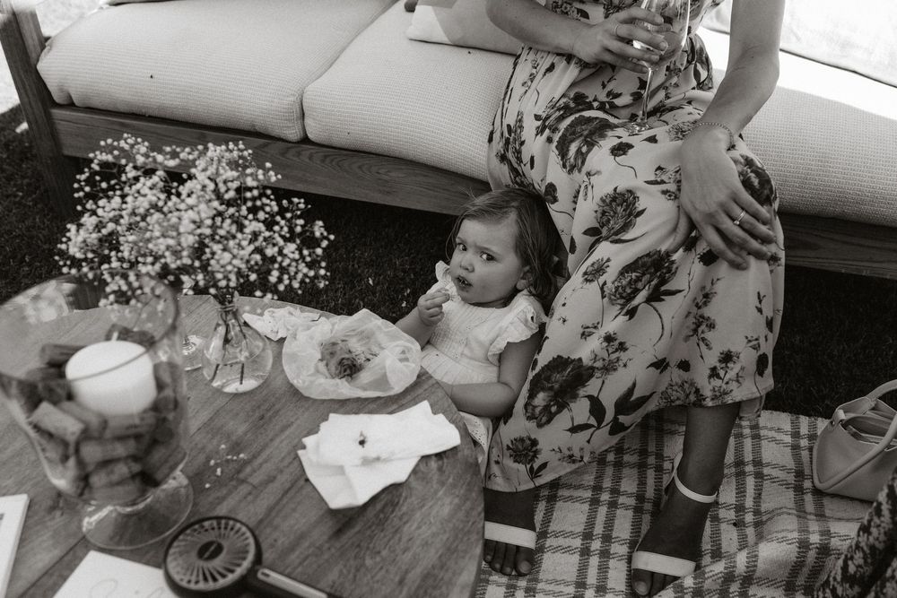 Black and white photo of a baby sitting beside an adult in a floral dress near a wooden table with flowers.