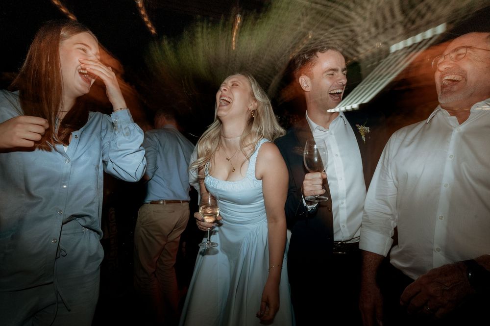 A group of joyful wedding guests laughing and dancing together on a dimly lit dance floor at a reception celebration.