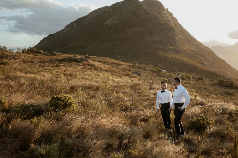 Couple walking along a scenic mountain trail during golden hour with dramatic clouds and rolling hills in the background.