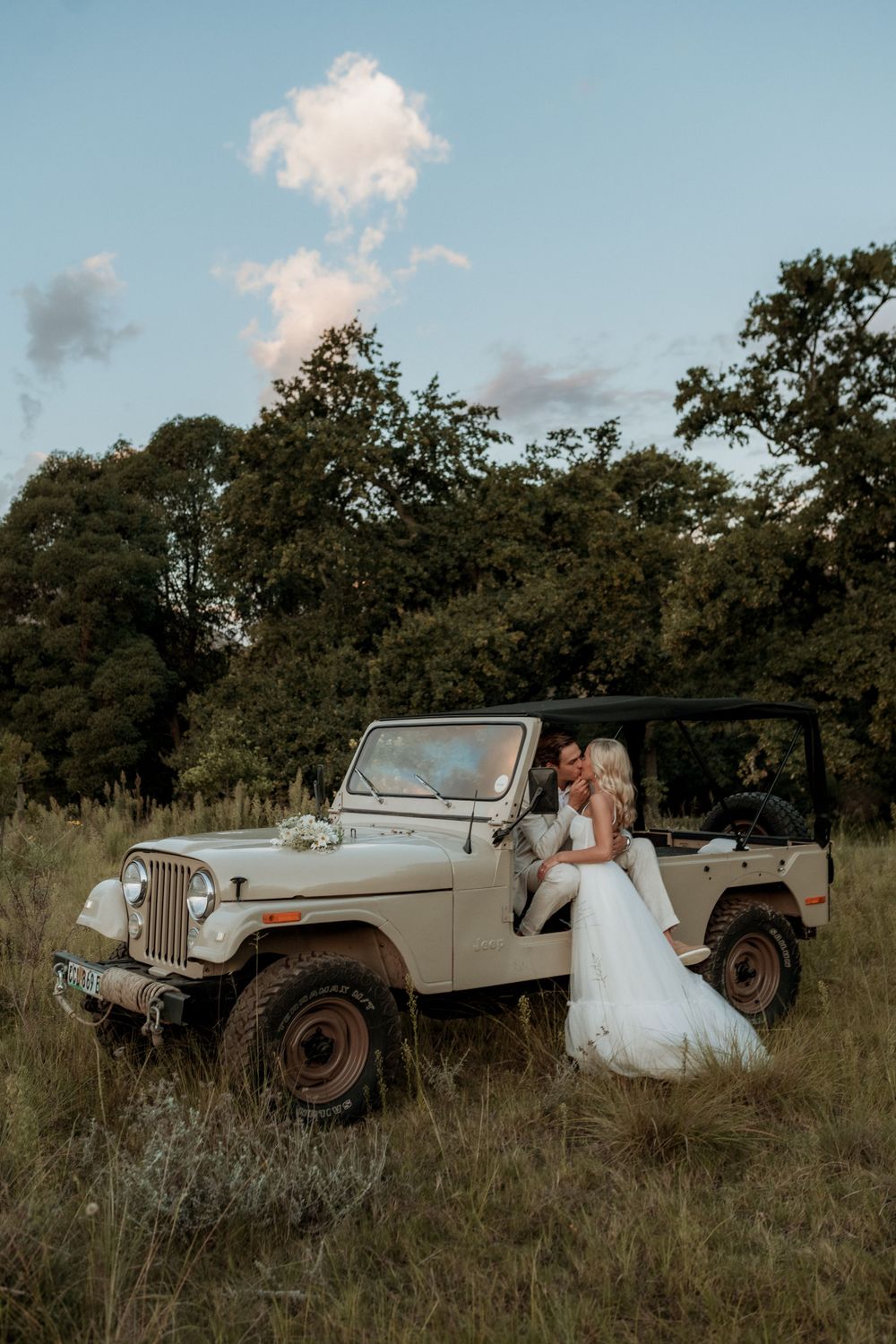 Vintage beige Jeep parked in tall grass field against backdrop of trees and blue sky with white clouds at sunset.