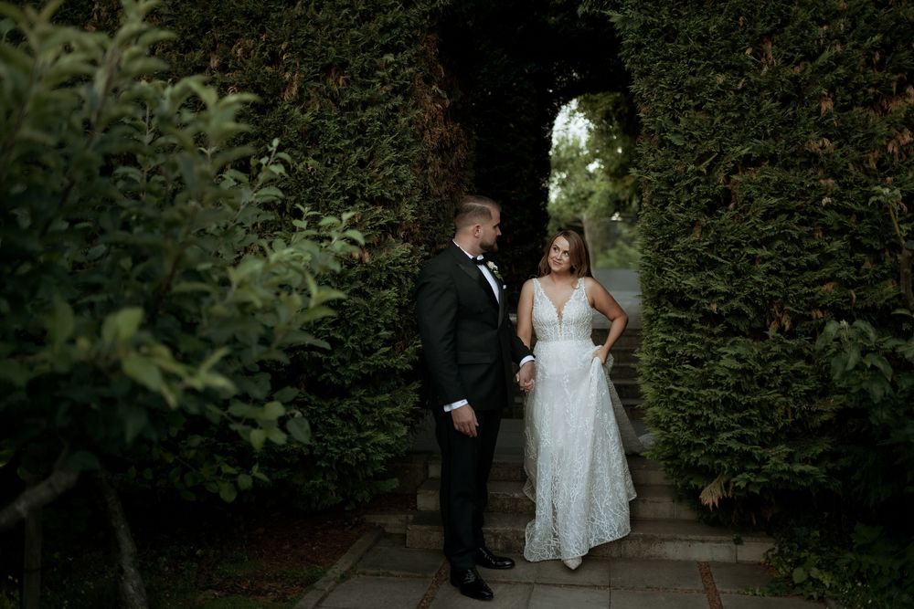 Couple walks through lush garden pathway during outdoor wedding photoshoot.