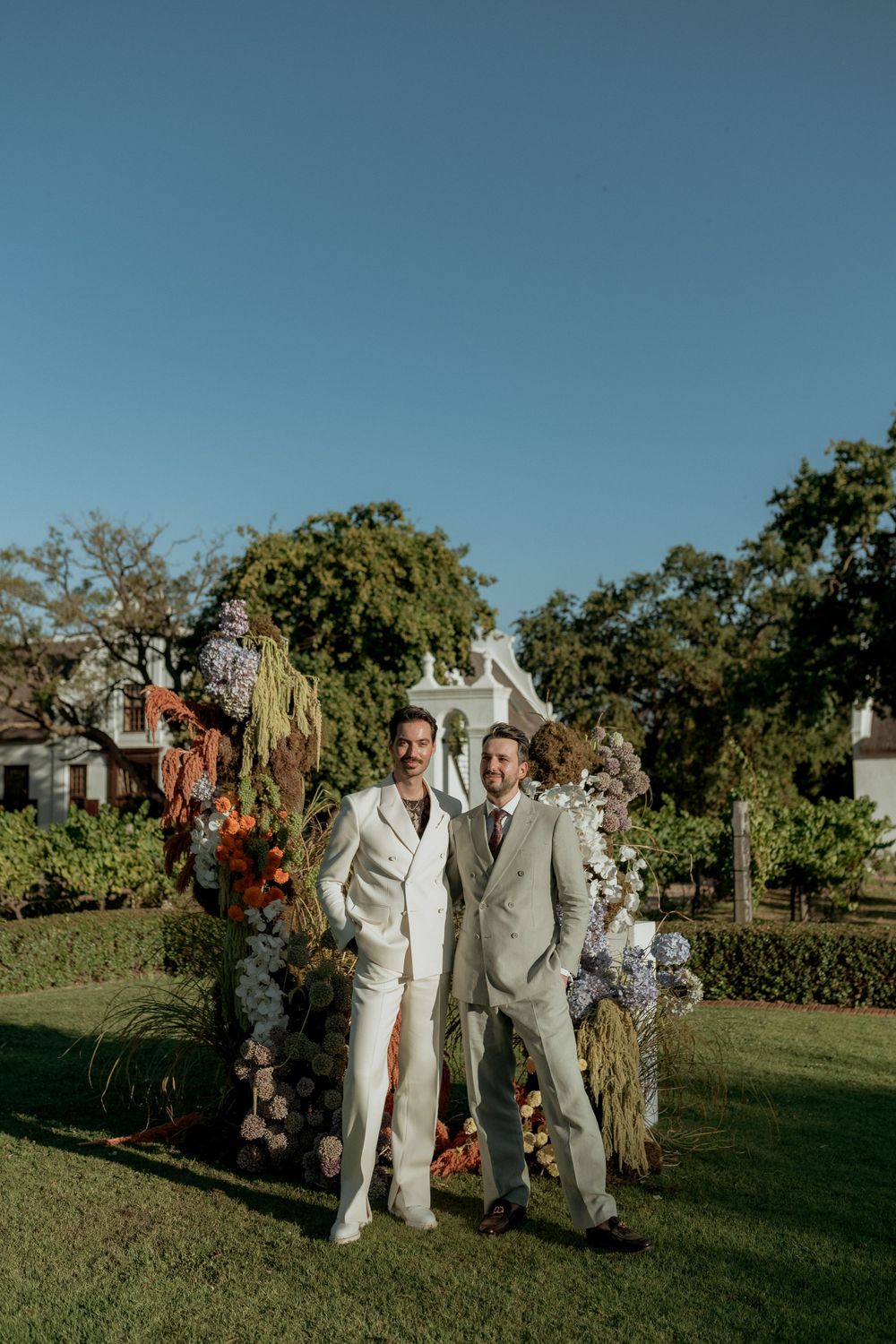Two people in light colored suits pose together in a garden setting with floral arrangements.