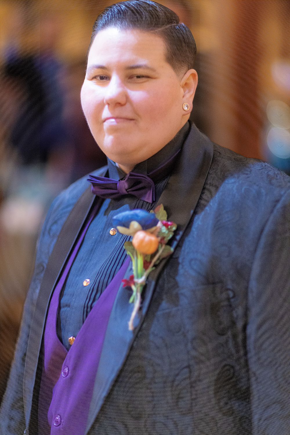 Wedding guest in formal attire wearing a black suit with purple vest and orange boutonniere.