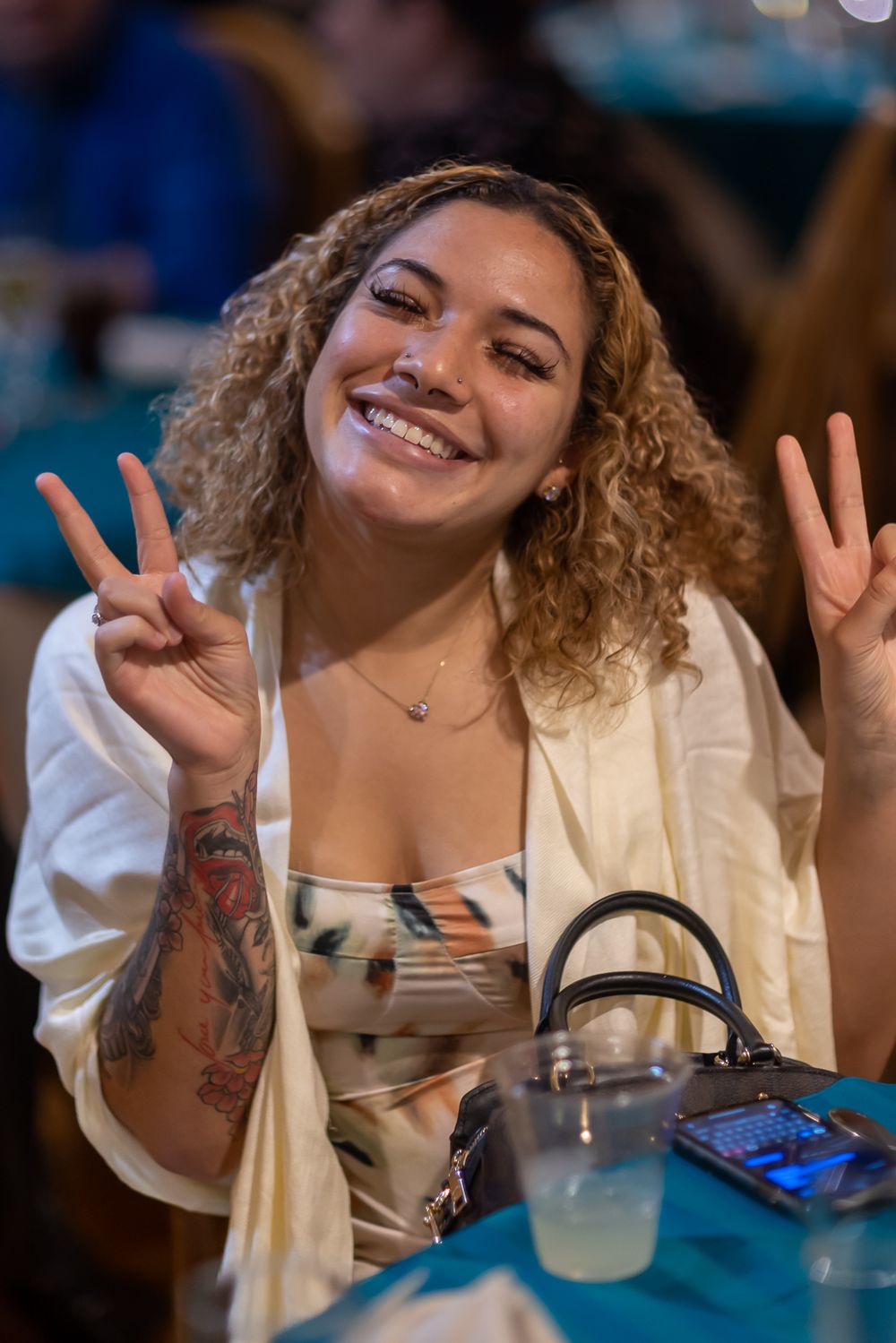 Person with curly hair and white top smiling and making peace signs at a restaurant table.