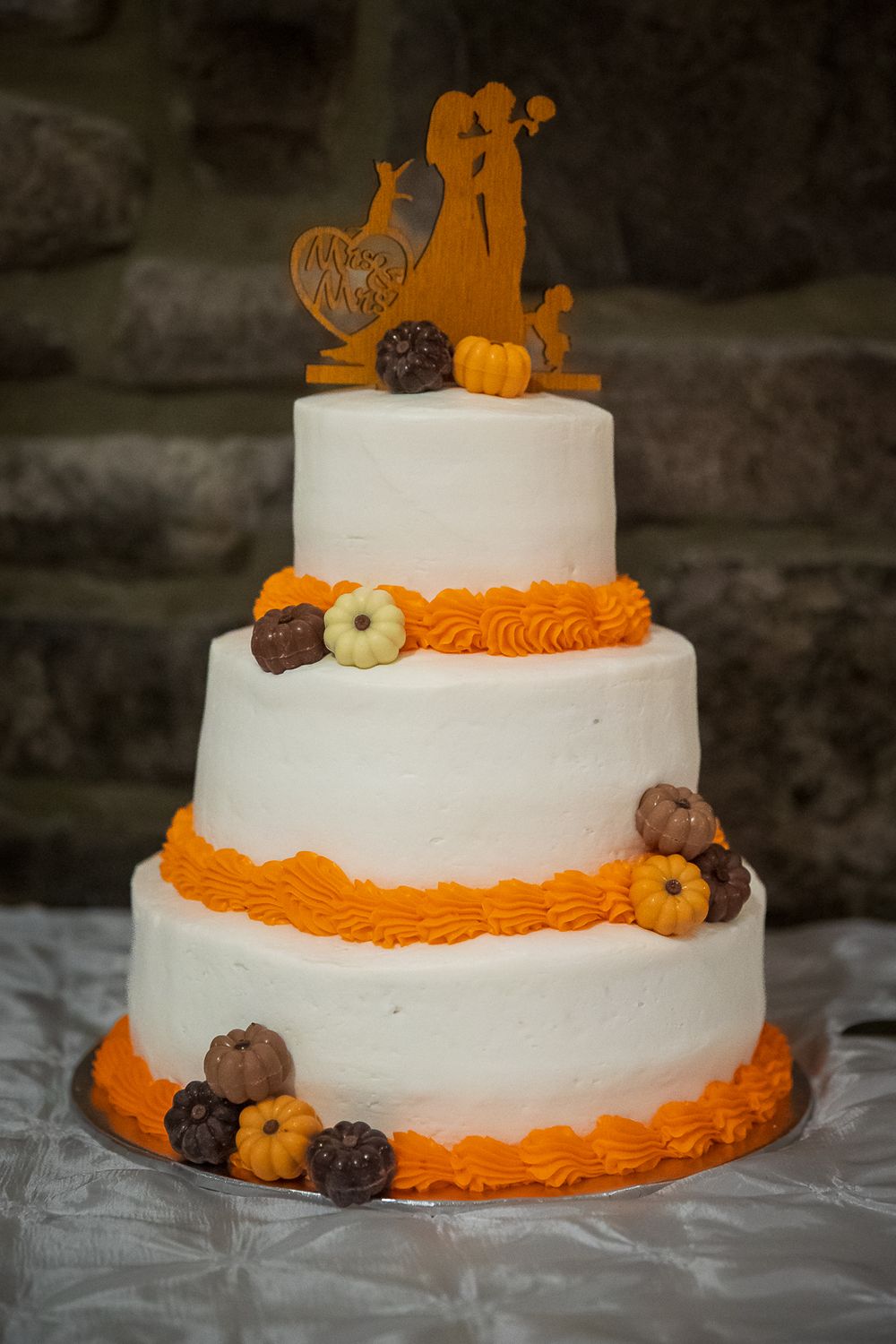 A white tiered cake decorated with orange trim and fall-themed toppers featuring pumpkins and autumn designs.