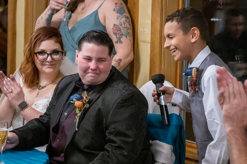 A wedding party celebrates during a reception with guests clapping and cheering in an elegant indoor venue.