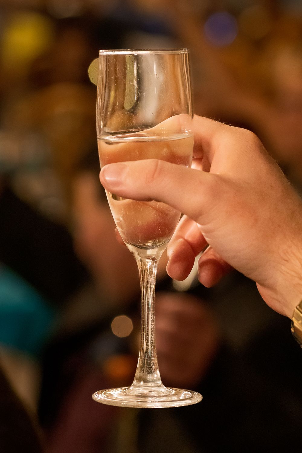 A hand holds up an elegant champagne flute with sparkling wine against a warm bokeh background.
