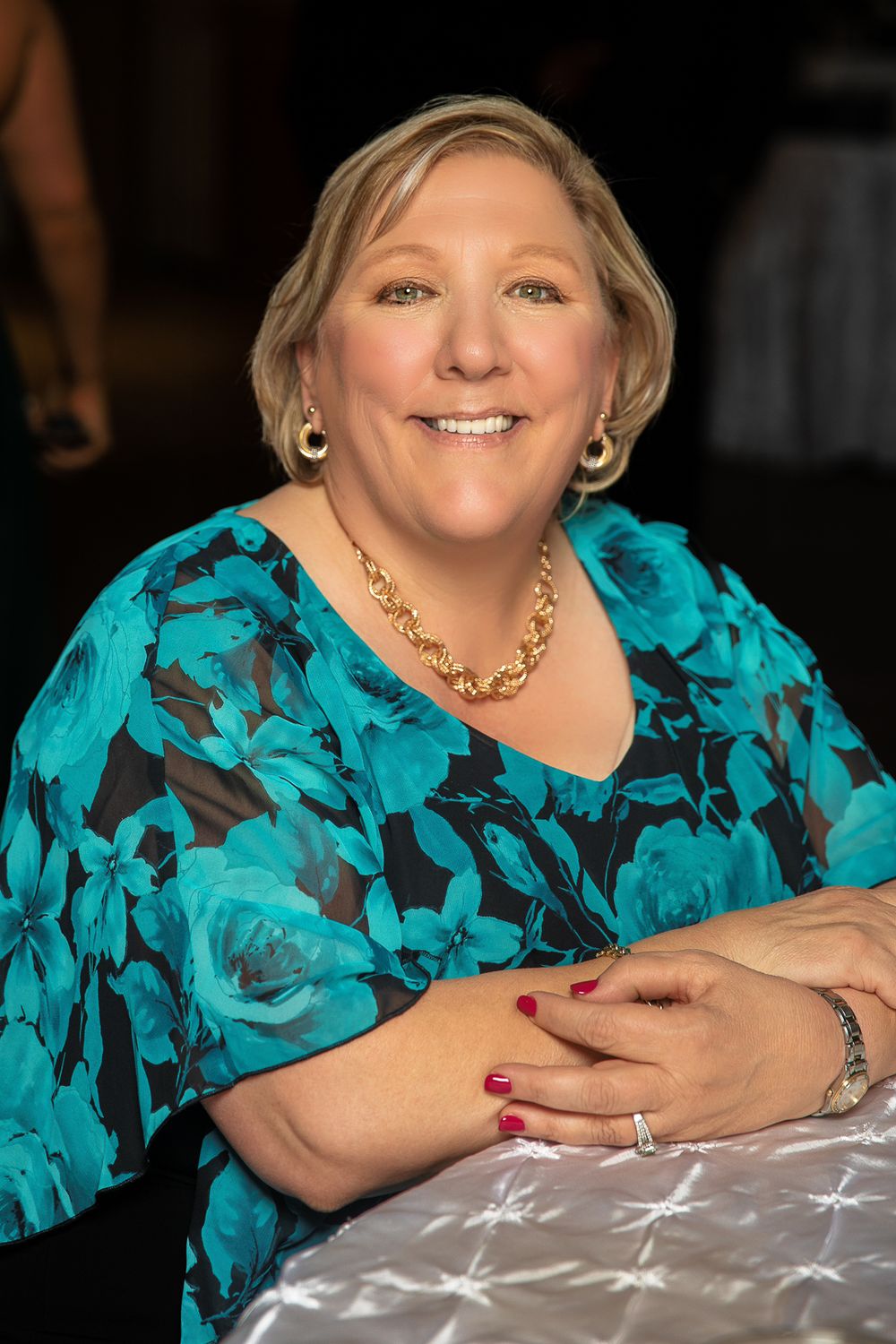 A person wearing a vibrant turquoise and black floral print top smiles warmly while sitting at a table.