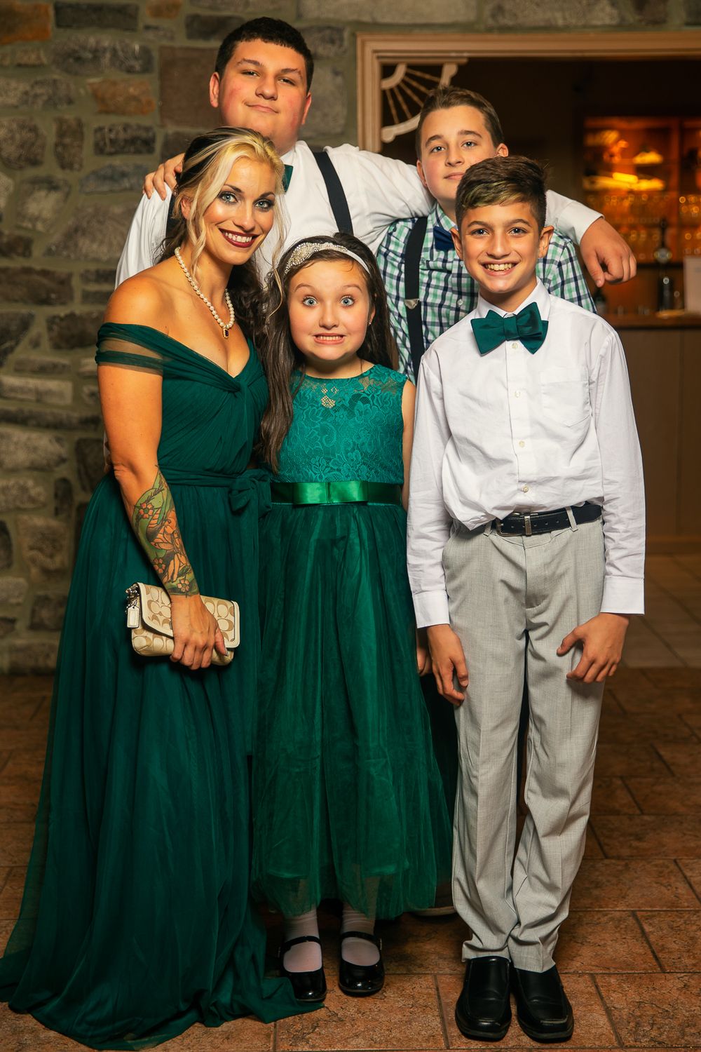 A family dressed in formal attire poses together, with emerald green dresses and dress clothes against a rustic stone wall.