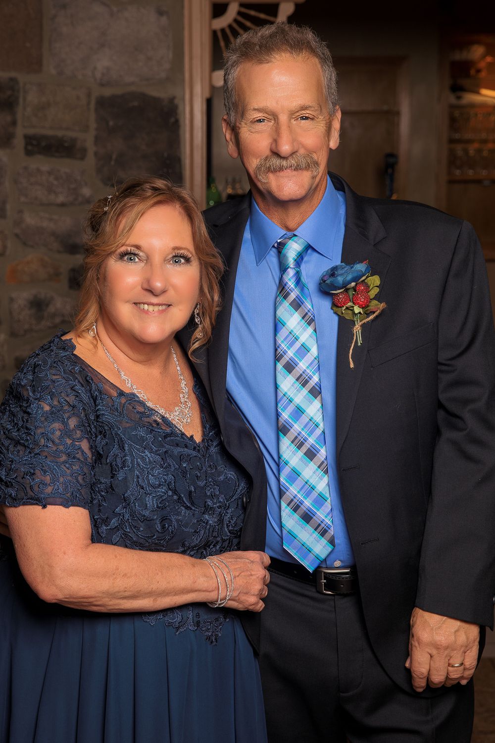 A couple dressed formally in navy blue attire at an evening event, with the man wearing a blue tie and boutonniere.