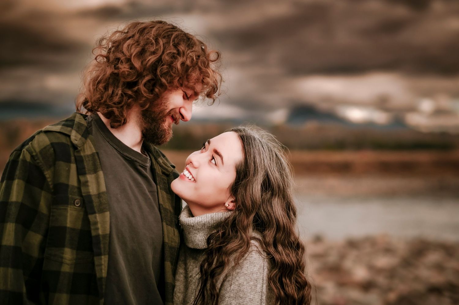 A couple shares a tender moment outdoors against a moody landscape backdrop while wearing cozy sweaters and plaid.