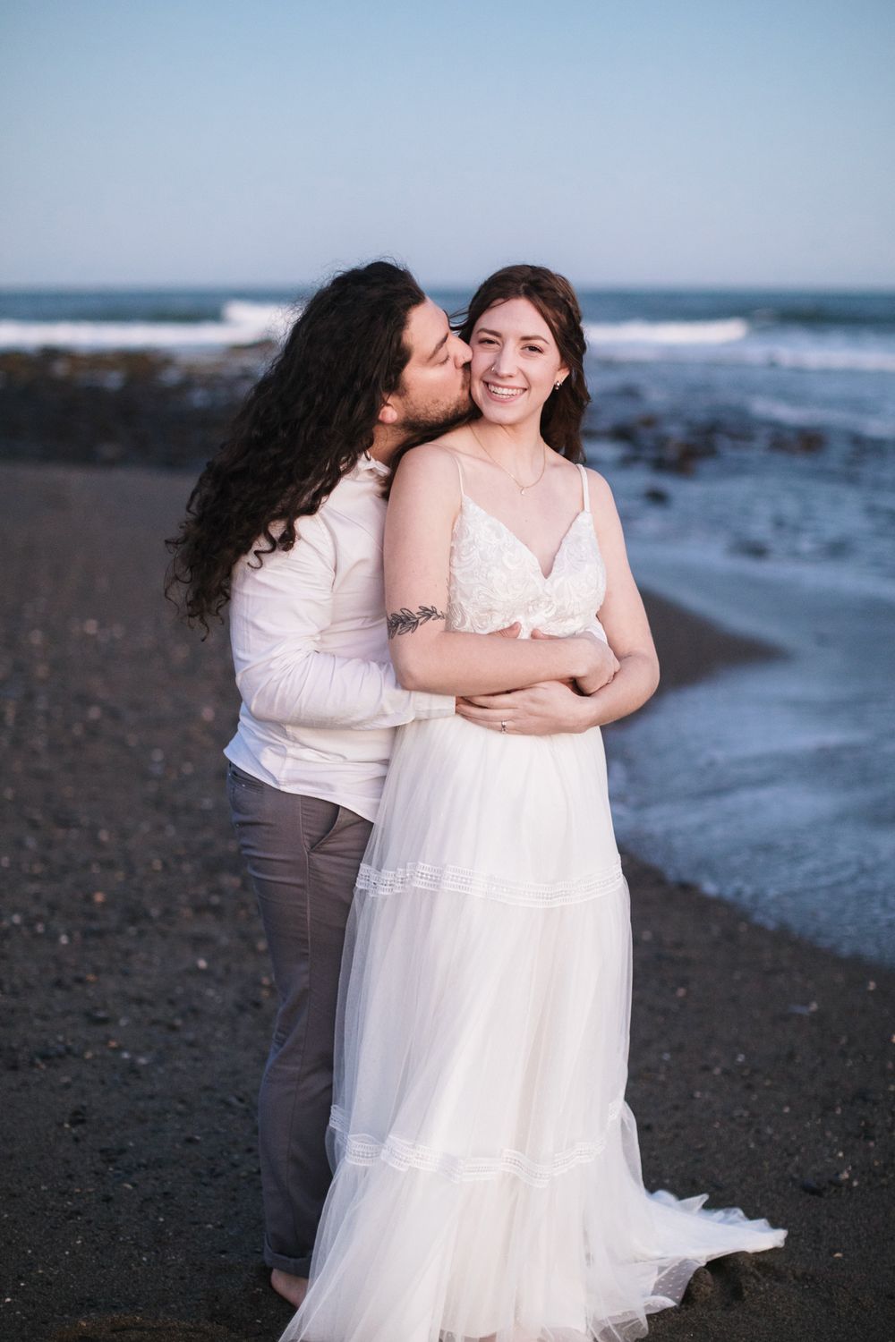 Husband kisses wife on cheek wearing wedding attire on dark sand beach in Rye, New Hampshire. Seacoast NH wedding photographer