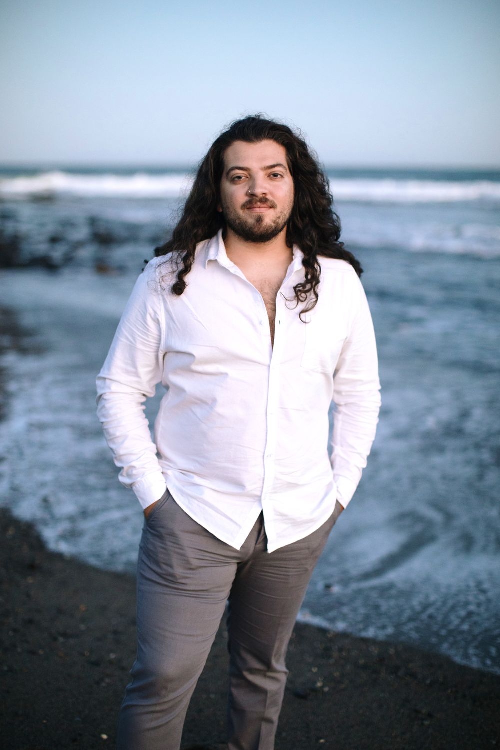 Husband with long, dark wavy hair poses with hands in his pockets wearing a white colored shirt and gray pants on beach at blue hour In Rye.