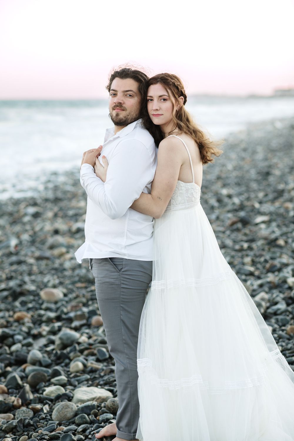 Woman in wedding dress embraces husband from behind standing on a rocky beach in North Hampton New Hampshire. Acadia wedding photographer.