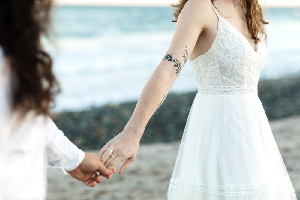A close up view of woman in wedding dress with tattoo on arm holding husband's hand with rolling waves at North Hampton Beach NH.