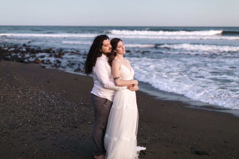 A wife leans back on her husband standing on a dark sand beach in Rye, Seacoast New Hampshire gazing out at ocean together at blue hour.