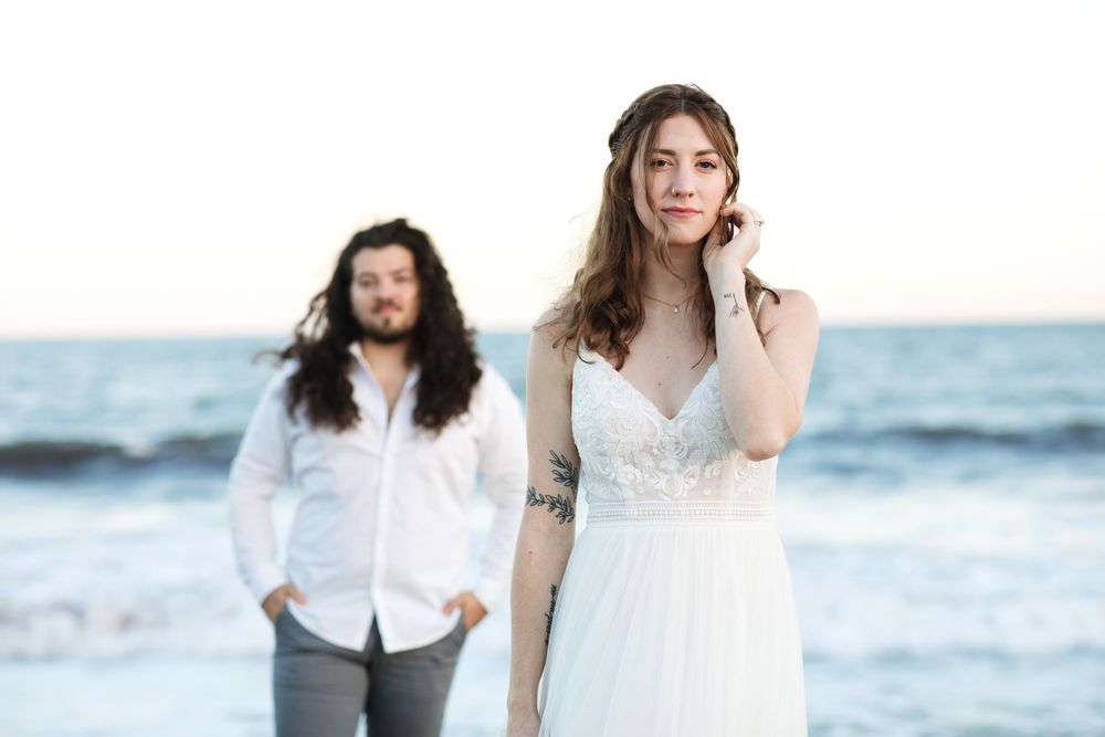 Bride holds her hair looking into camera with husband in background wearing wedding attire at North Hampton Beach, New Hampshire.