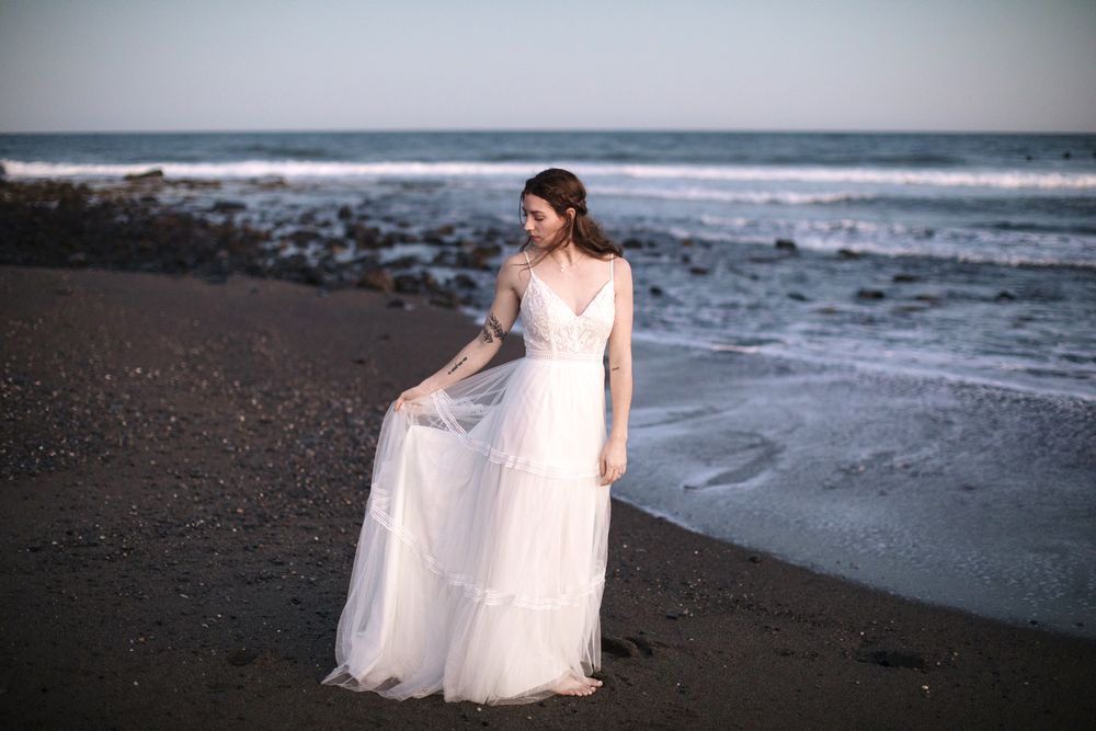 Woman in bridal gown gazes down at dress on a dark sand beach at dusk in Rye, New Hampshire. Rye, Hampton York wedding photographer