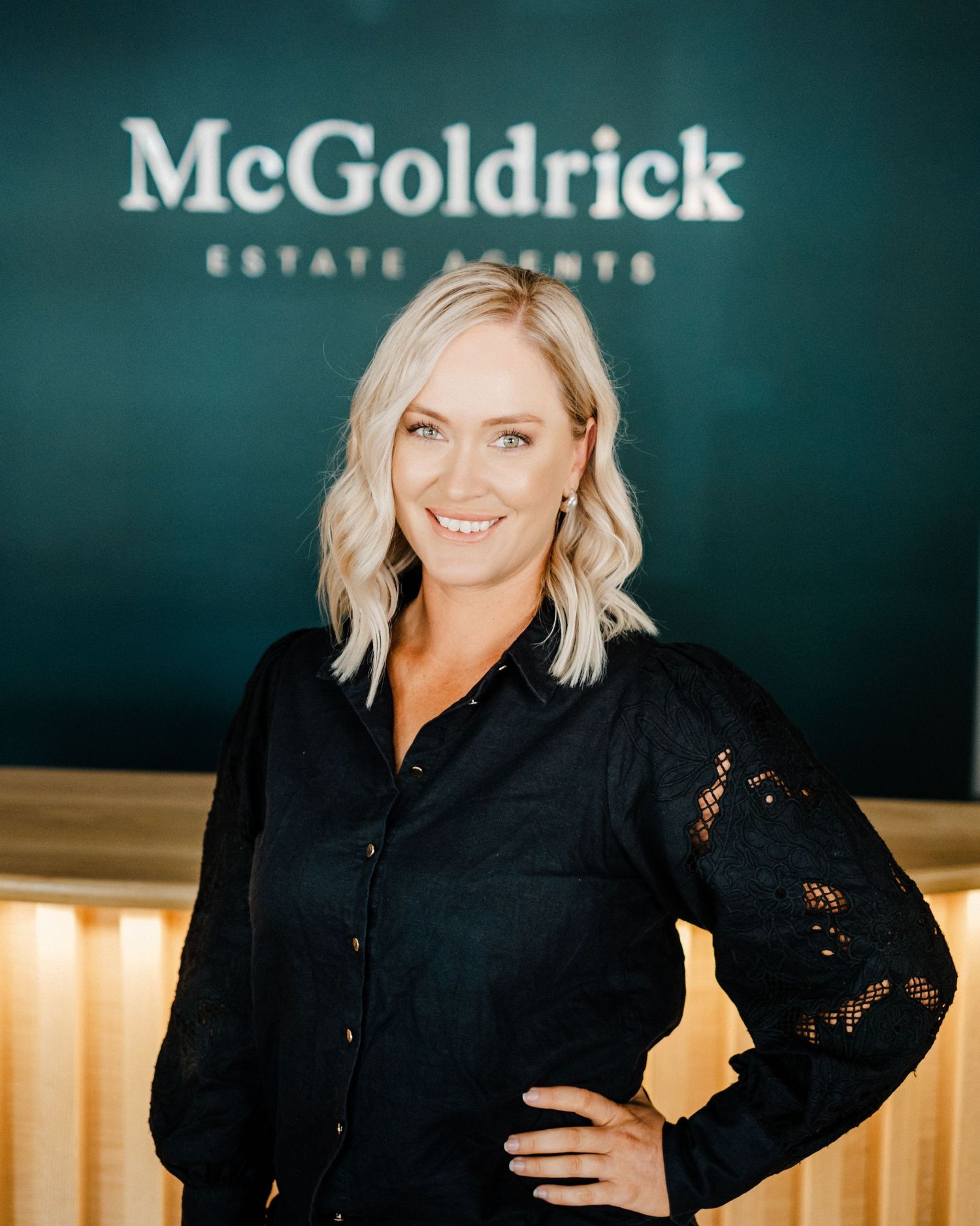 Professional real estate agent poses in front of McGoldrick company logo wall in black button-down blouse.