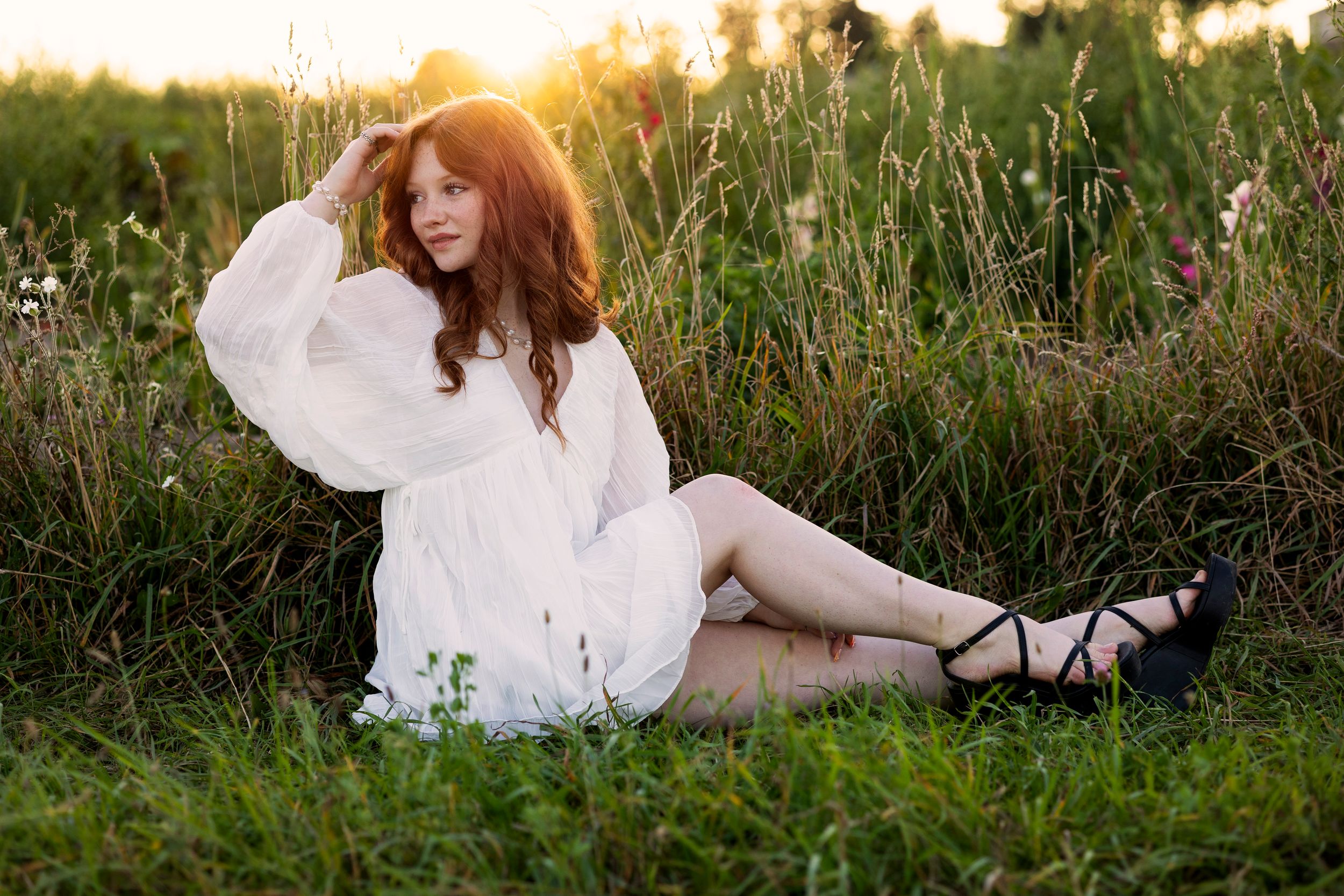 A senior in a flowing white dress reclines peacefully in a sunlit meadow during golden hour.
