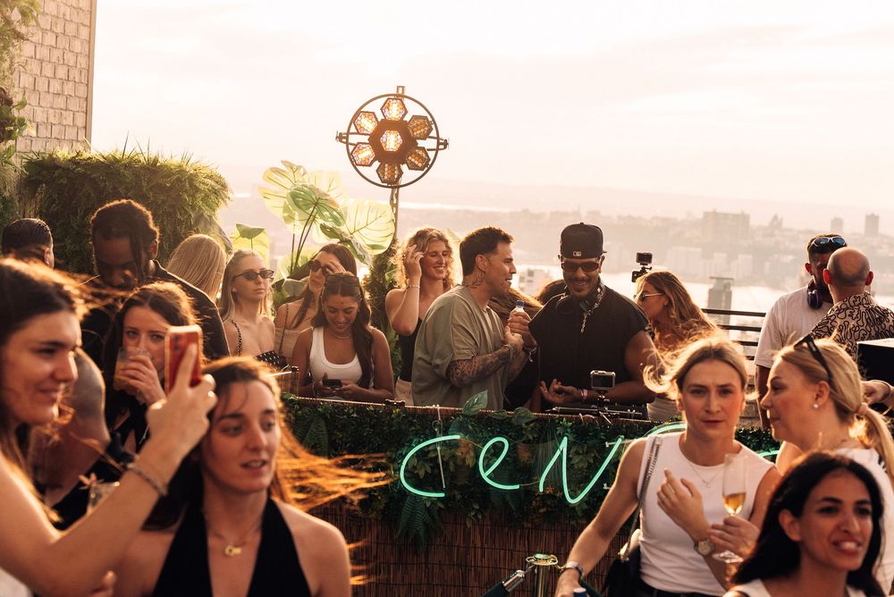 A crowd of people enjoying a rooftop party at sunset with a decorative metal flower sculpture in the background.