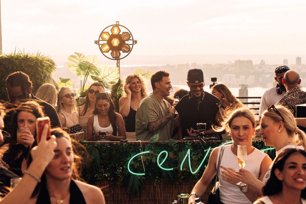A crowd of people enjoying drinks and socializing at an outdoor rooftop party during sunset.
