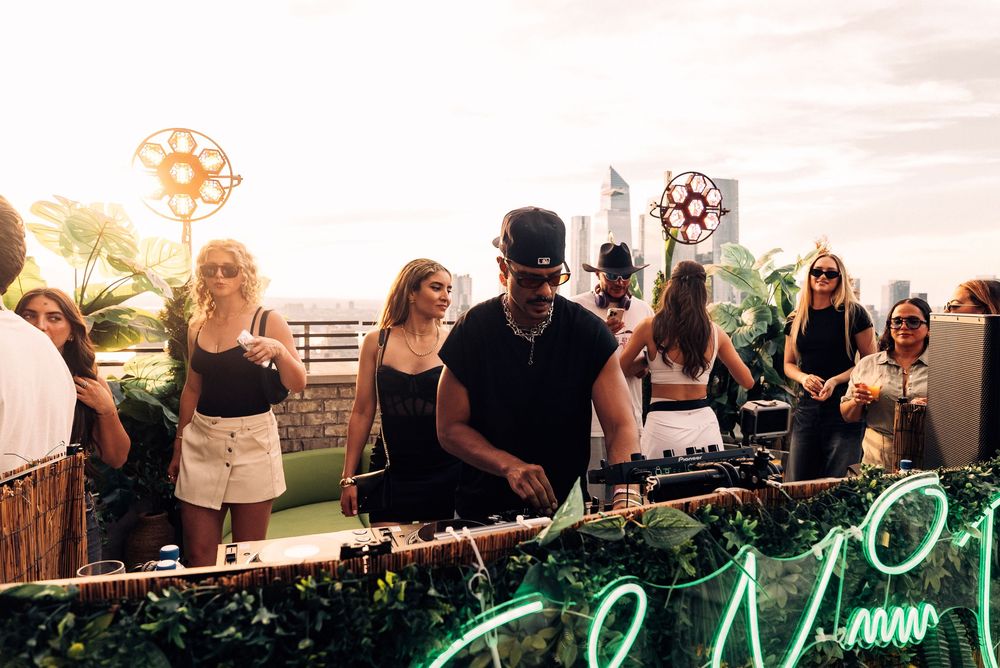 DJ performs at an outdoor rooftop party with neon signage and crowd of people enjoying the sunset atmosphere.