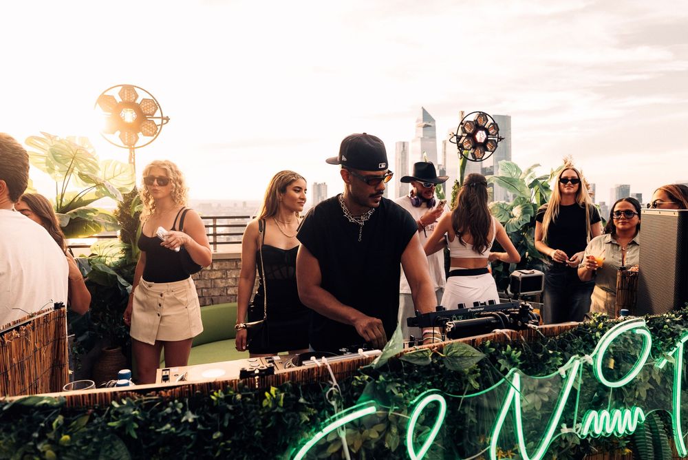 Outdoor party scene with DJs performing on turntables at sunset with neon lights and palm trees in background.