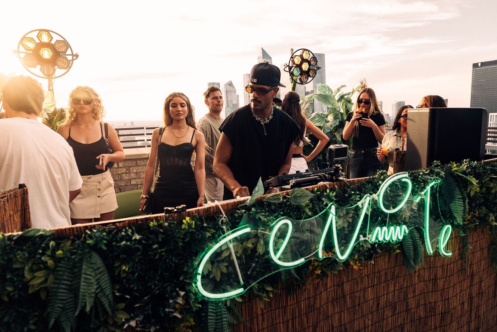DJ performs at an outdoor party with a glowing neon sign and greenery-decorated booth during sunset.