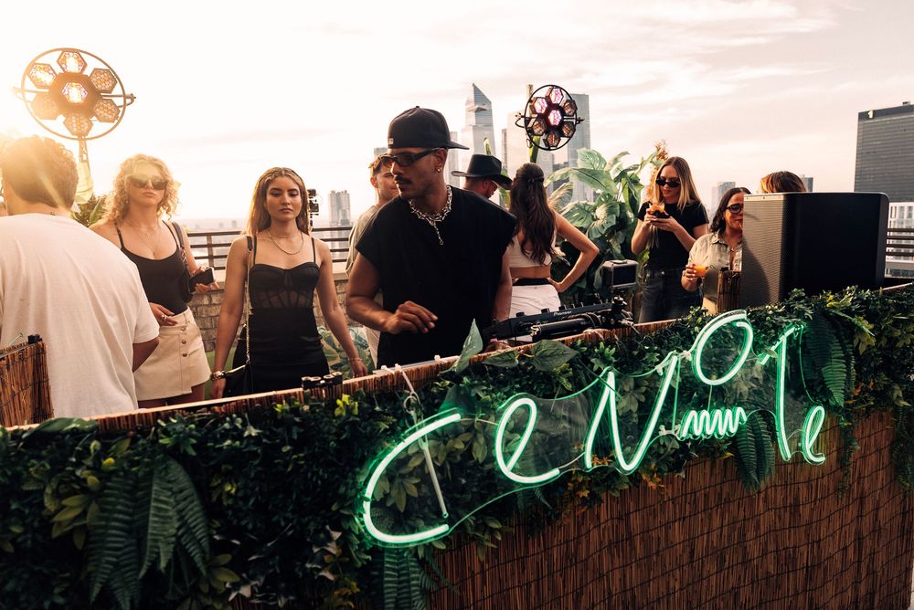 Guests mingle at a rooftop party with a glowing neon sign in the background during sunset.