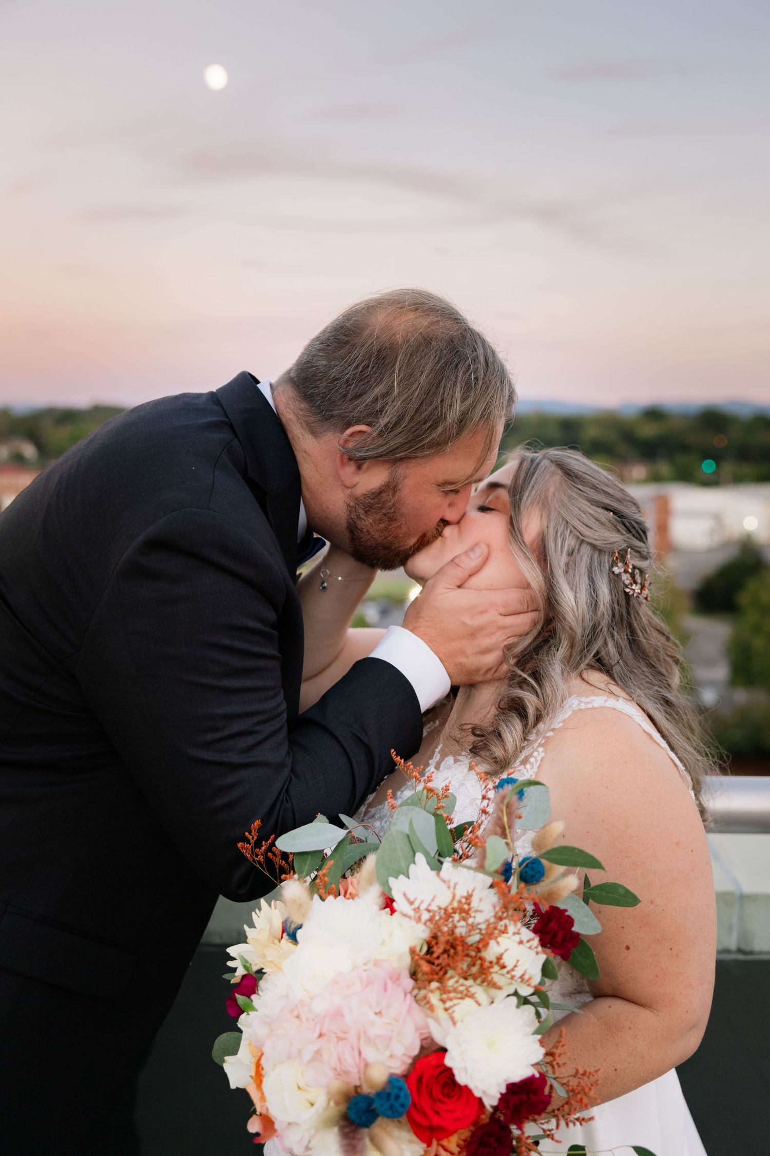 A romantic sunset wedding couple shares a kiss on a rooftop while holding a colorful bouquet of flowers.