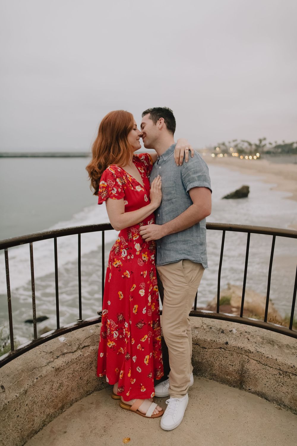 A couple in a romantic embrace on a seaside balcony, woman in red floral dress, man in gray shirt, with ocean views.