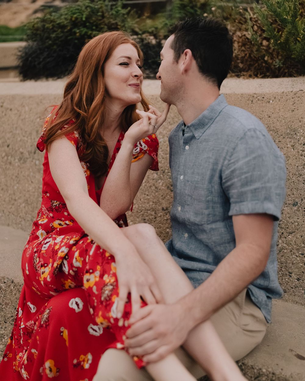 Redhead woman in red floral dress sitting with man in blue shirt on stone steps outdoors.