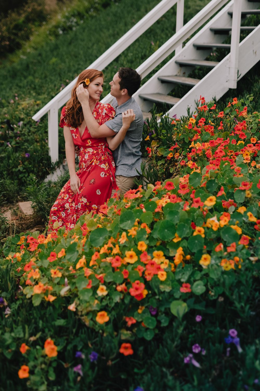 Couple embracing near white staircase surrounded by vibrant orange and red flowers in a lush garden.