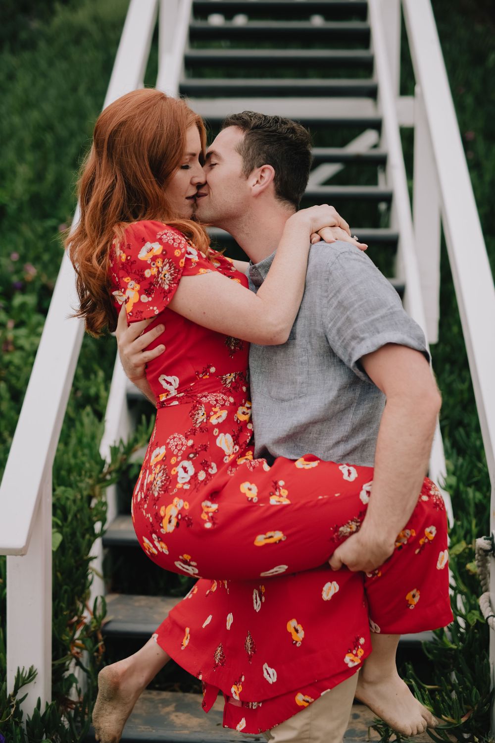 Couple sharing a romantic kiss on white garden stairs, woman in red floral dress lifted by man in gray tee.