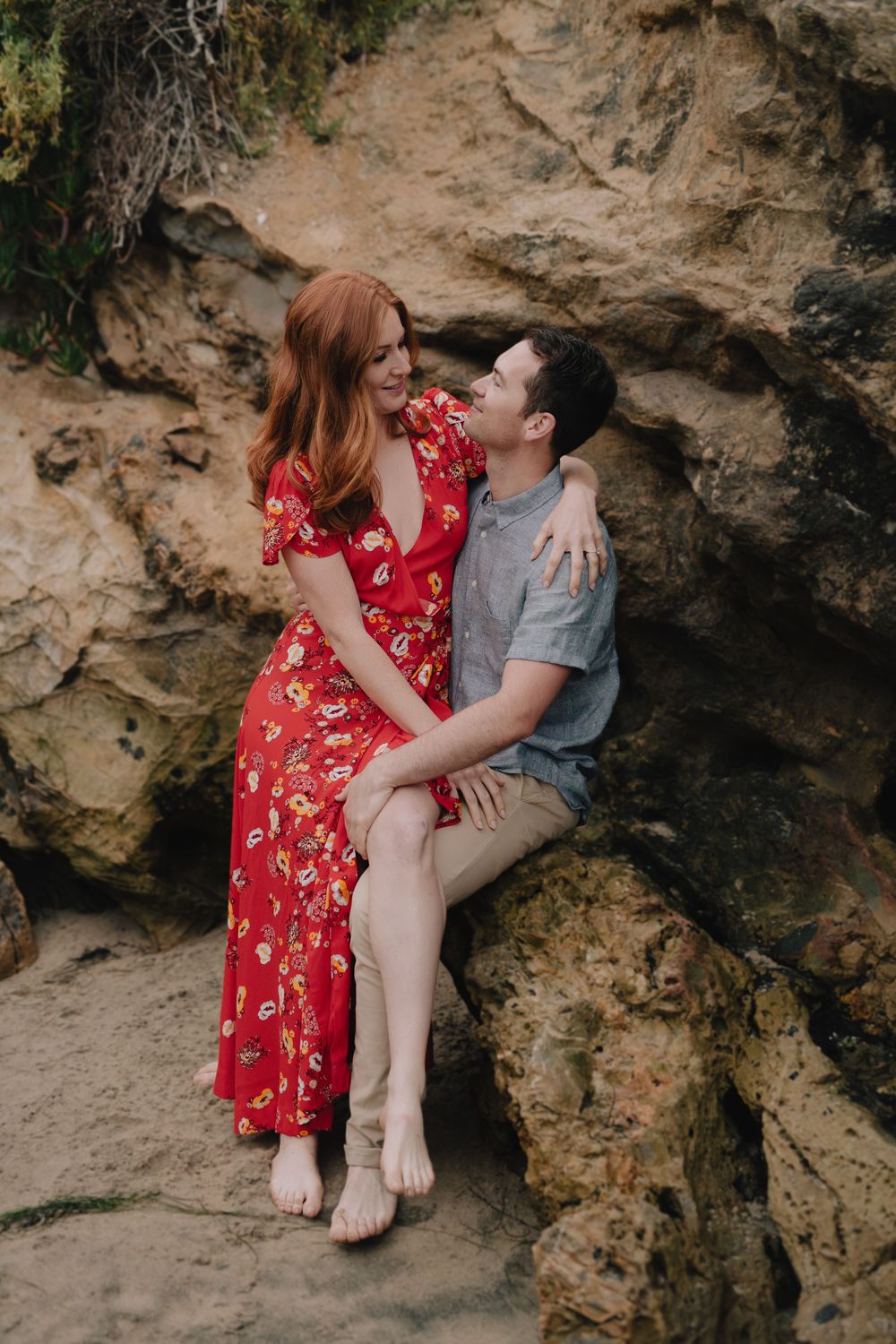 A couple sits on coastal rocks, woman in red floral dress and man in gray shirt, sharing a romantic moment.